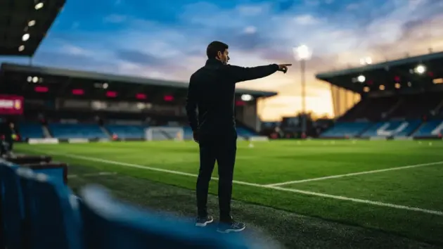Football coach on touchline with blue and claret stadium atmosphere at golden hour