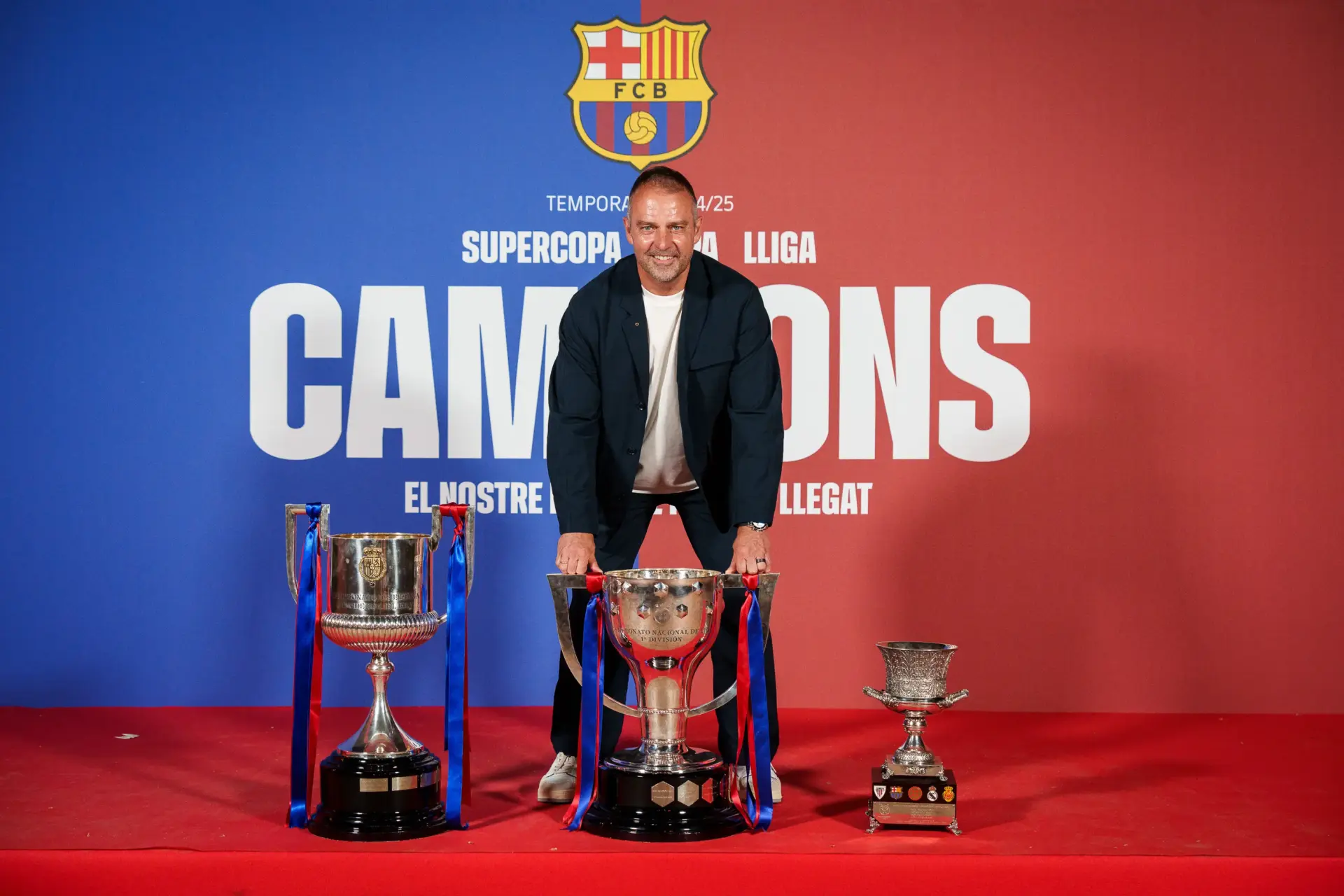 Hansi Flick poses with three football trophies against a Barcelona backdrop.