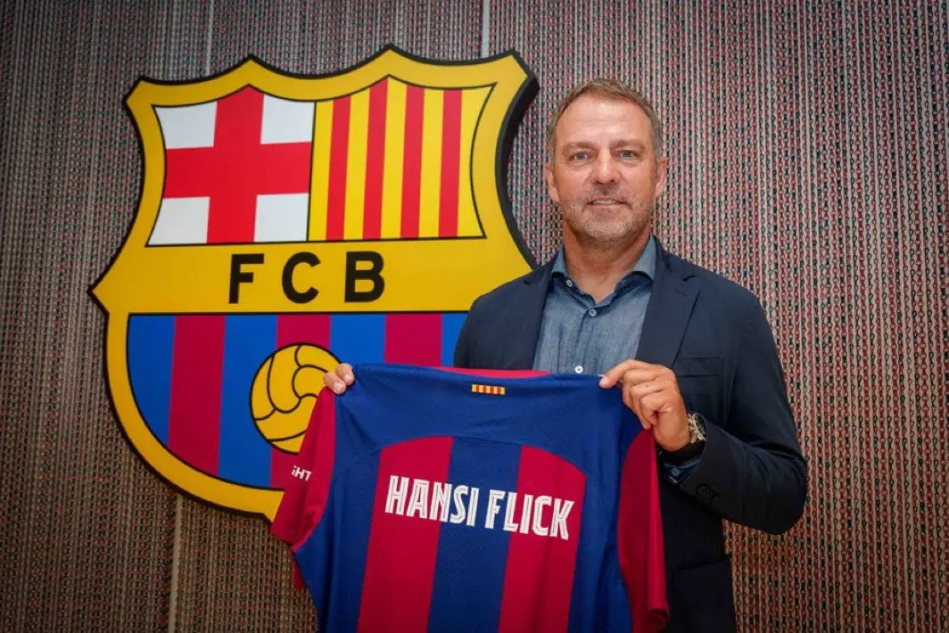 Hansi Flick holding an FC Barcelona jersey with his name on it in front of the club crest
