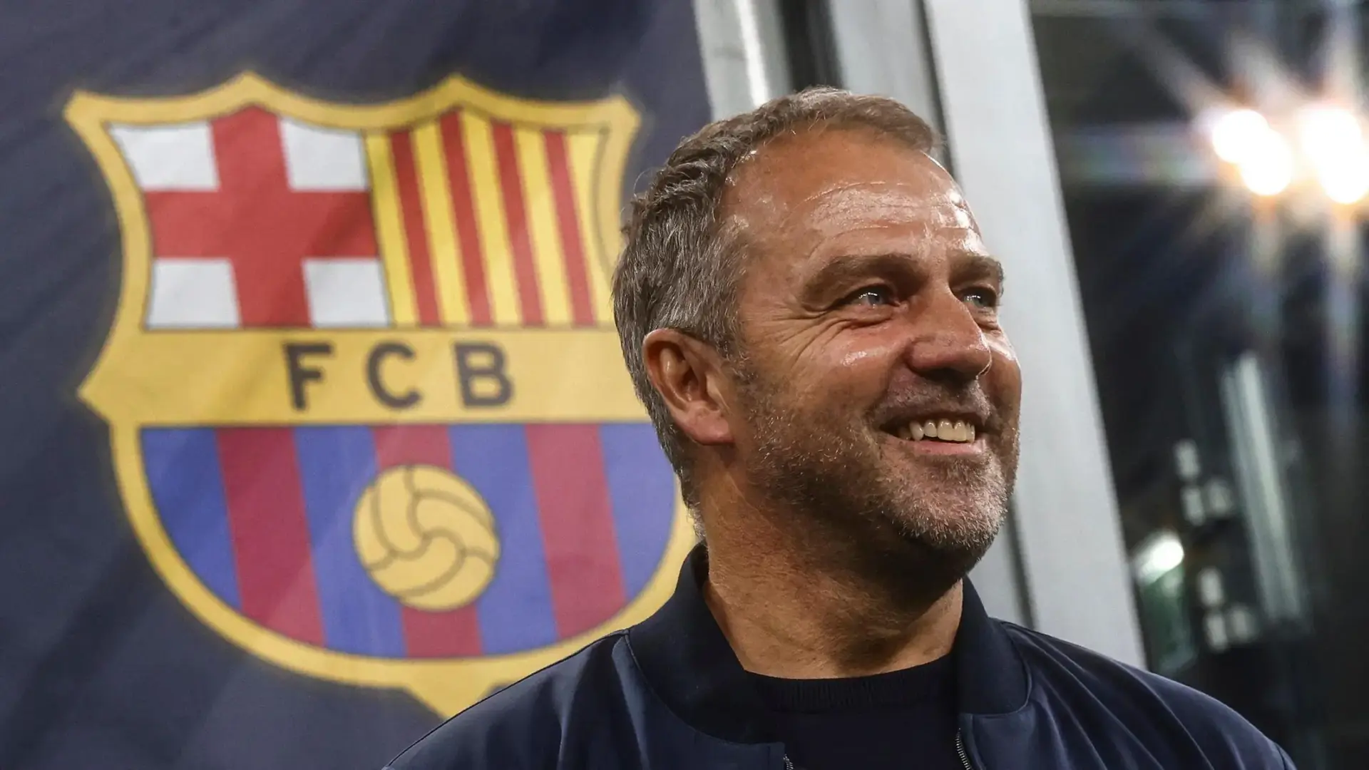 Hansi Flick smiling in front of FC Barcelona's logo.