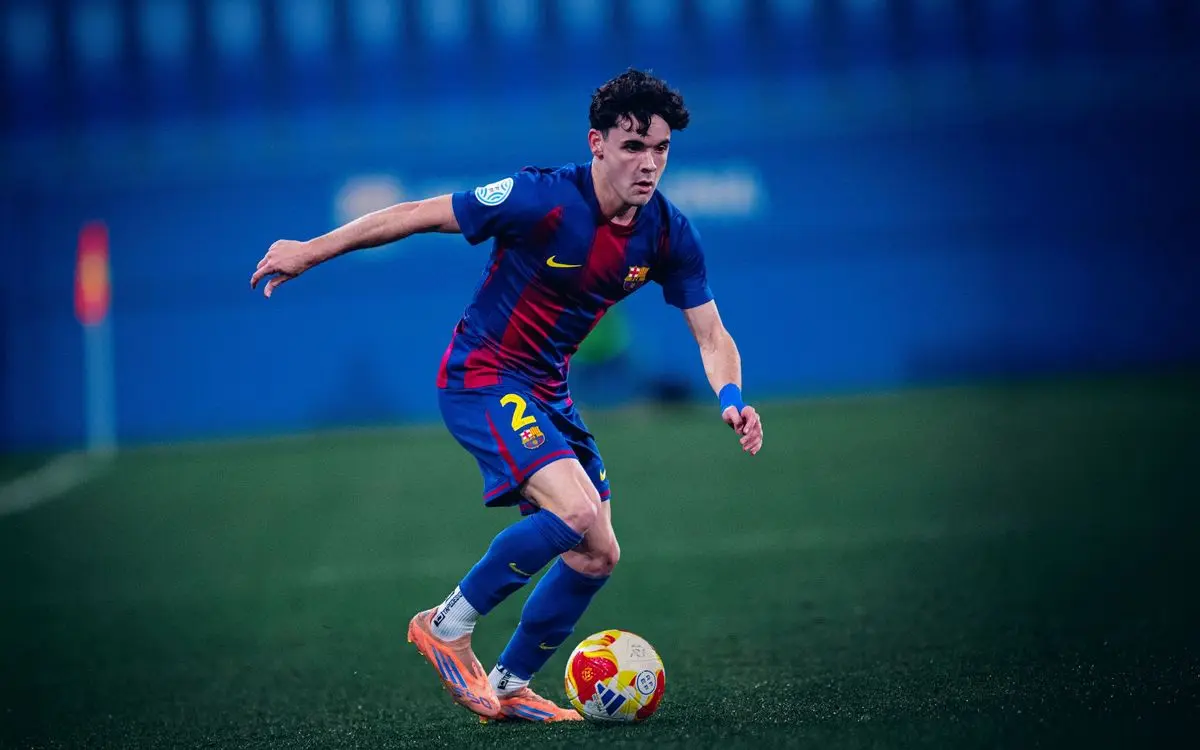 Joan Anaya in a FC Barcelona Athletic blue and red kit dribbling a soccer ball on a green pitch.