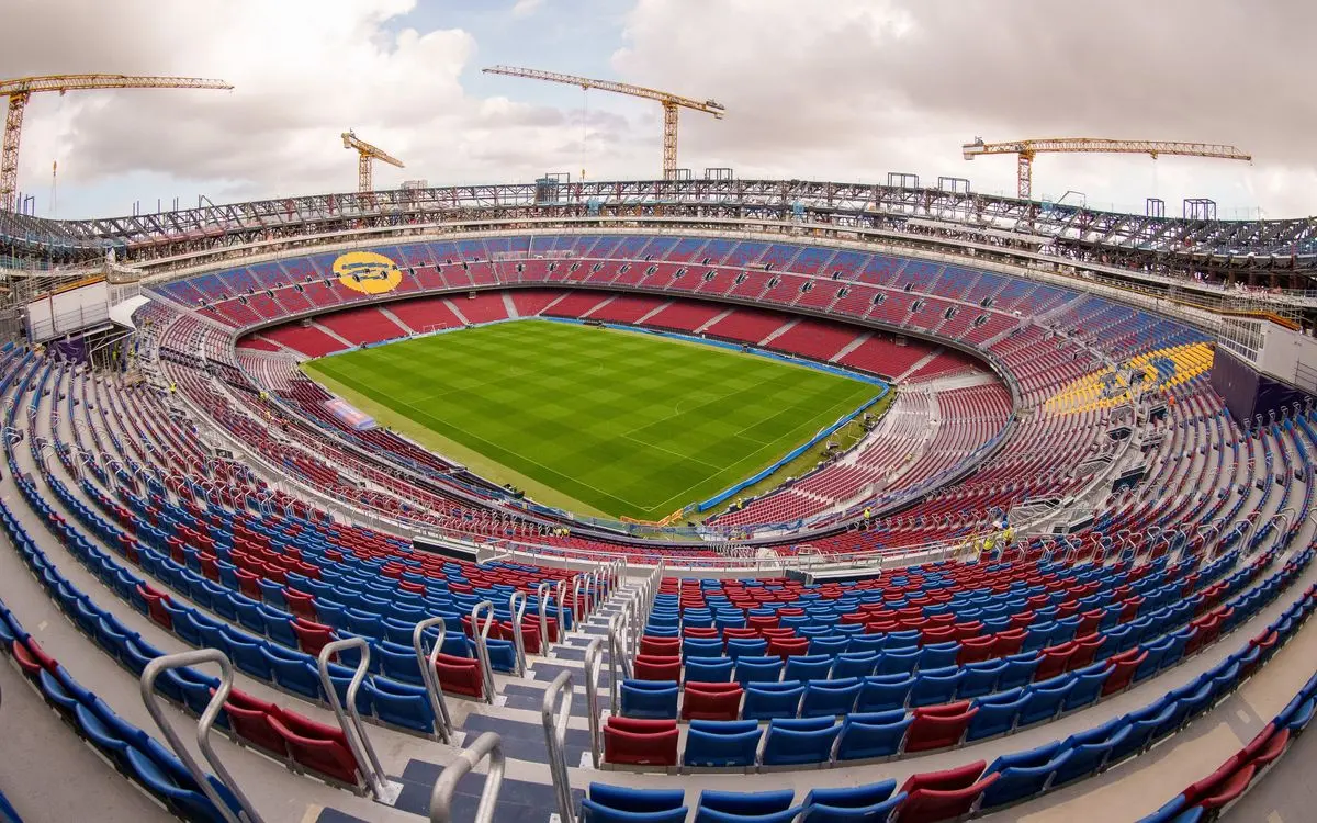 Wide-angle view of the Camp Nou stadium in Barcelona with red and blue seats and construction cranes above.
