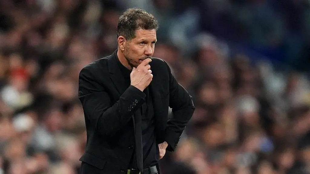 Diego Simeone wearing a black suit and tie, looking thoughtful on the sidelines of a football match.