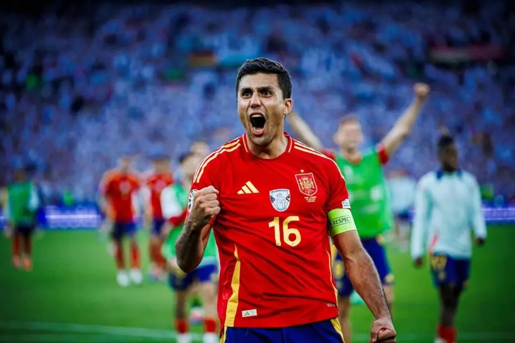 Rodri wearing a red Spain national team jersey with number 16, celebrating with a shout on the pitch.