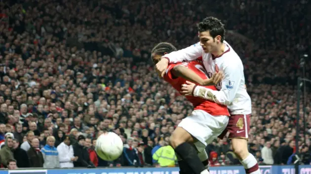 Cesc Fàbregas in a white kit during a football match, contending for the ball with another player.
